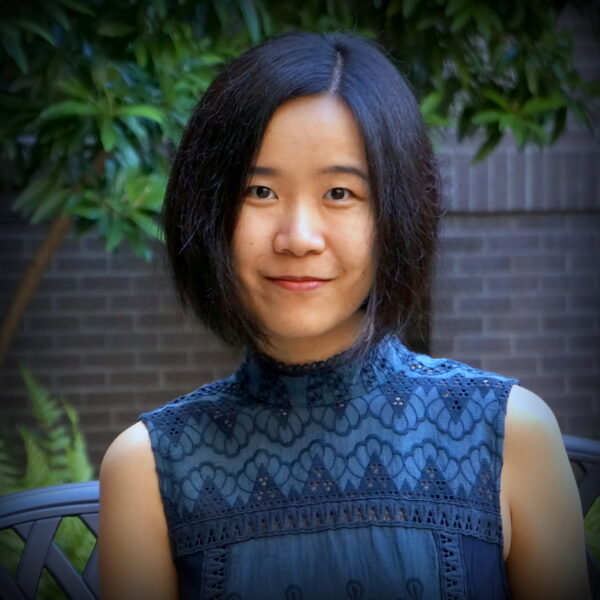 Profile photo of Dr. Yingchen He. She has dark hair and wears an indigo sleeveless mock neck top. There are green plants and brick walls in the background.